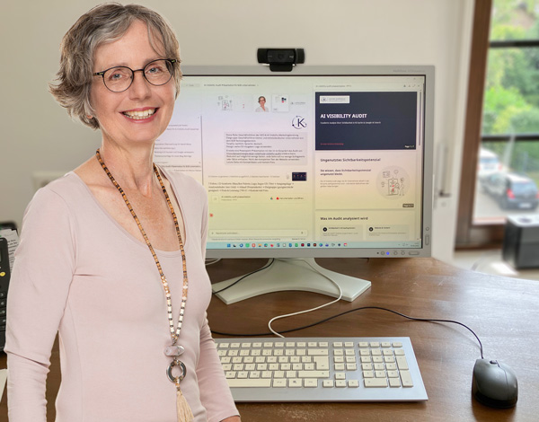Katrin Knörrich steht lachend vor ihrem Schreibtisch. Darauf eine Tastatur und ein Monitor in dem ein Bild von einem geöffneten KI-System mit Text- und Bildausgabe zu sehen ist.